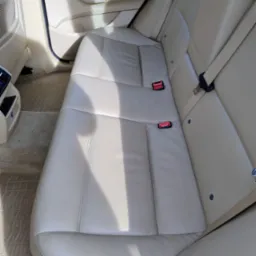 Beżowa tylna kanapa skórzana w samochodzie, widoczne pasy bezpieczeństwa i dywaniki podłogowe.