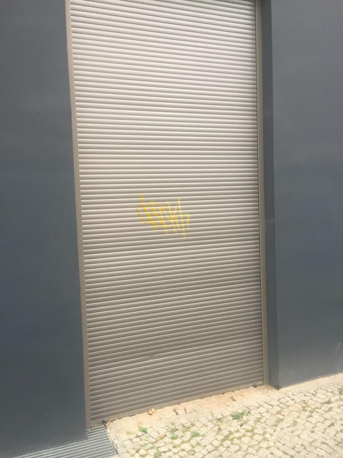 Zewnętrzna metalowa roleta garażowa z żółtymi graffiti na szarym tle ściany budynku.