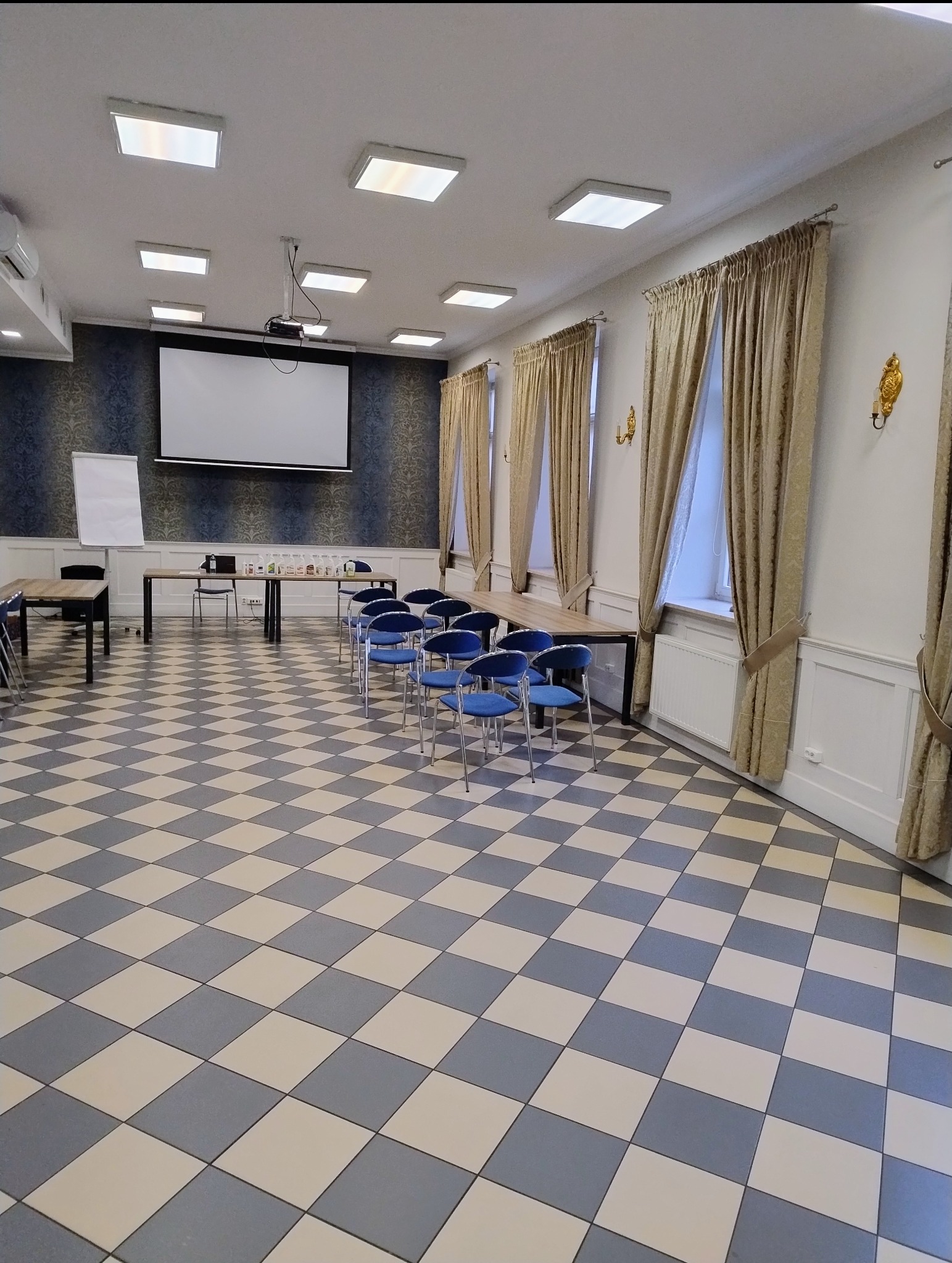 Sala konferencyjna z szachownicową podłogą, rzędem niebieskich krzeseł, stołem, ekranem projekcyjnym i flipchartem, przygotowana do prezentacji, widoczne środki czystości na stole.