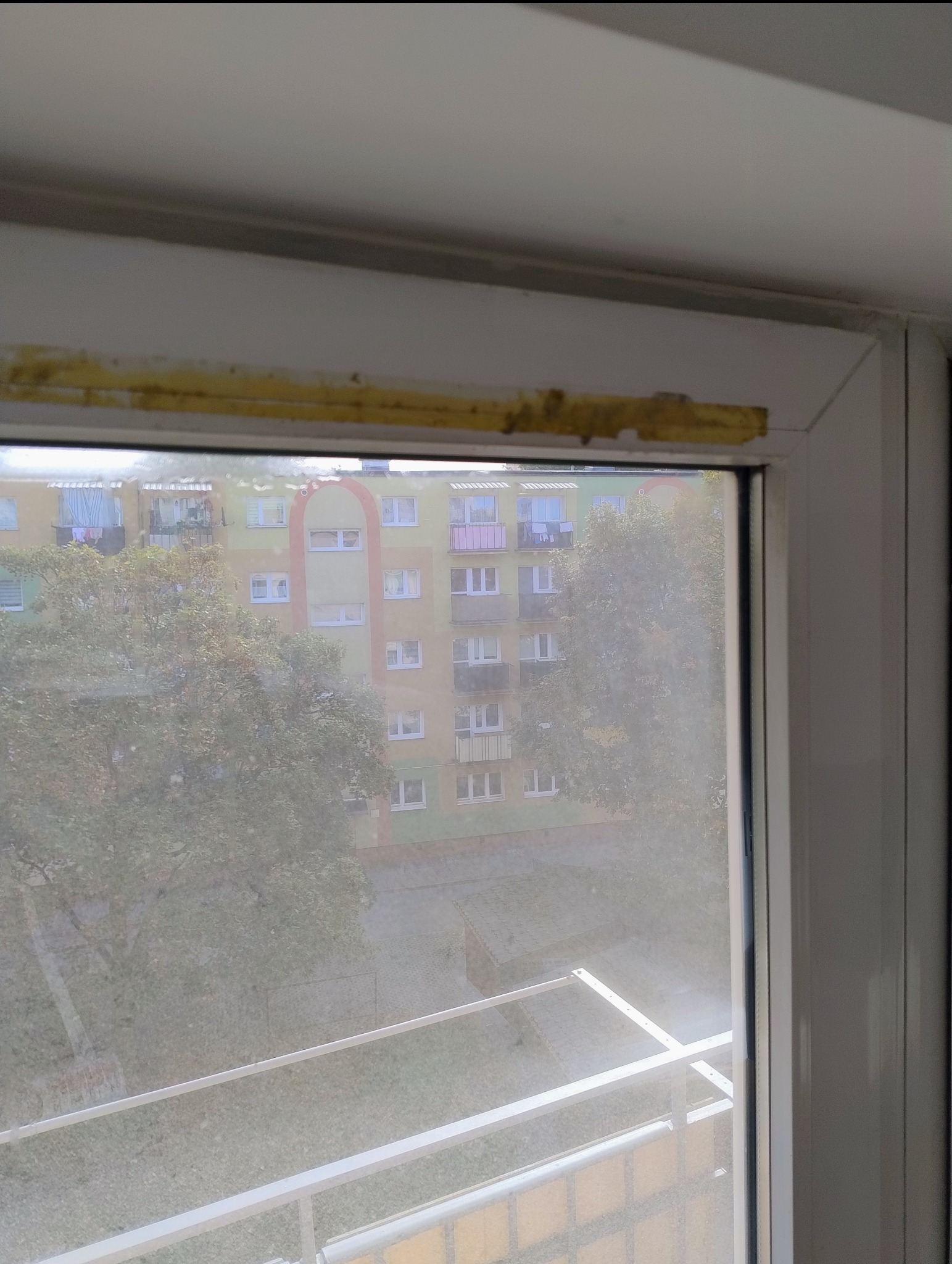 Brudne okno PCV z widokiem na blok mieszkalny i drzewa, widoczne ślady brudu na ramie okiennej.