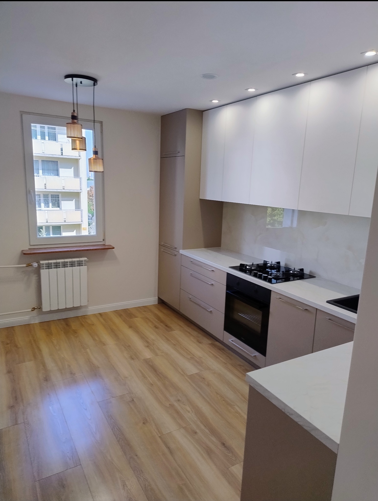 Nowoczesna, jasna kuchnia w Radomiu po remoncie, z drewnianą podłogą, beżowymi szafkami i białymi górnymi szafkami, widok na okno z grzejnikiem.