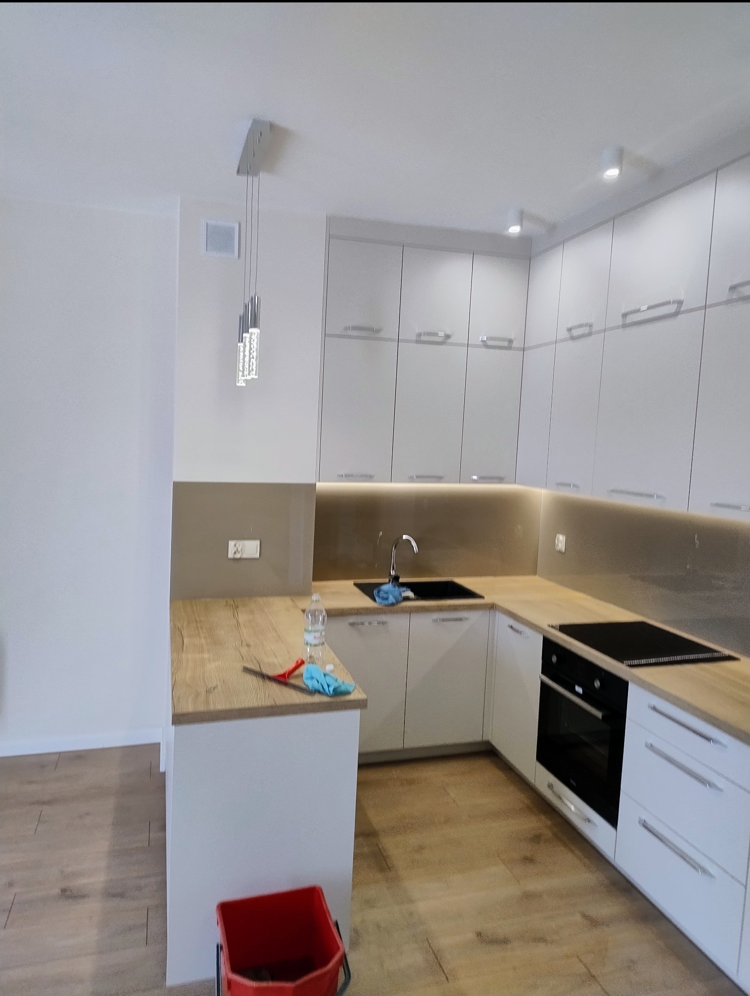 Wnętrze nowoczesnej, jasnej kuchni z białymi szafkami i drewnianym blatem; na blacie leży ściereczka i butelka płynu, a na podłodze stoi czerwone wiadro.