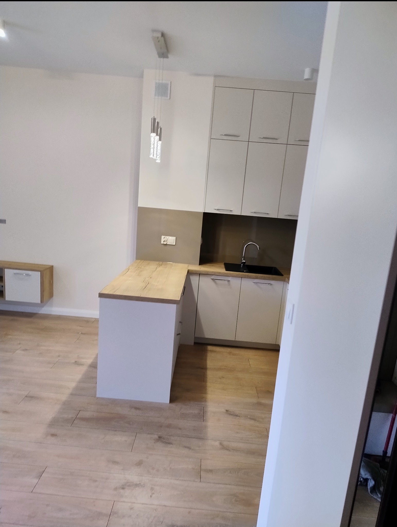 Wnętrze jasnej, nowocześnie urządzonej kuchni z drewnianym blatem i białymi szafkami, widok na minimalistyczny zlew i baterię, a także wiszące lampy kryształowe.