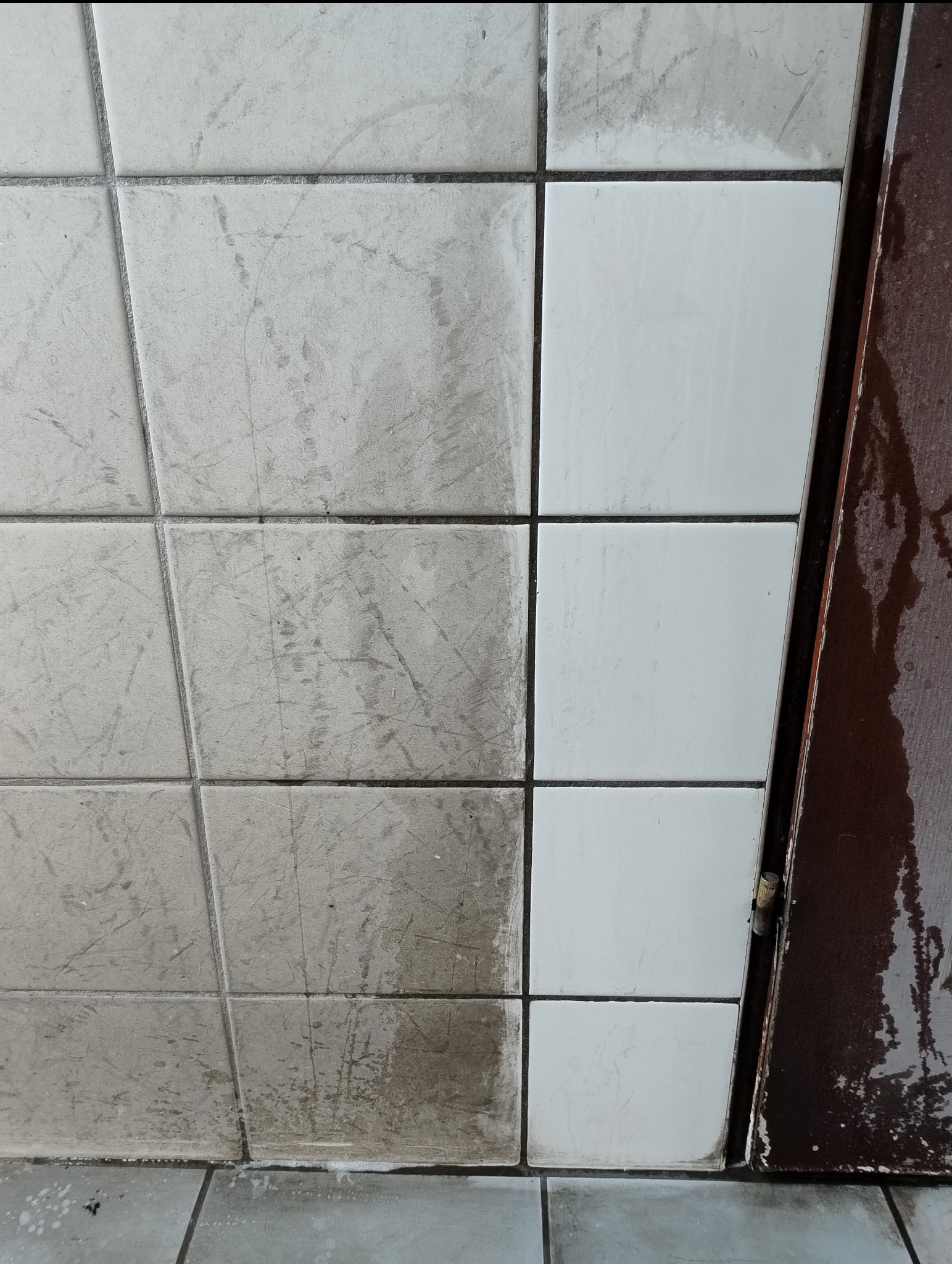 Porównanie stanu płytek ściennych przed i po czyszczeniu, widoczne zabrudzenia po lewej stronie i czysta powierzchnia po prawej, obok fragment zardzewiałej futryny drzwi.