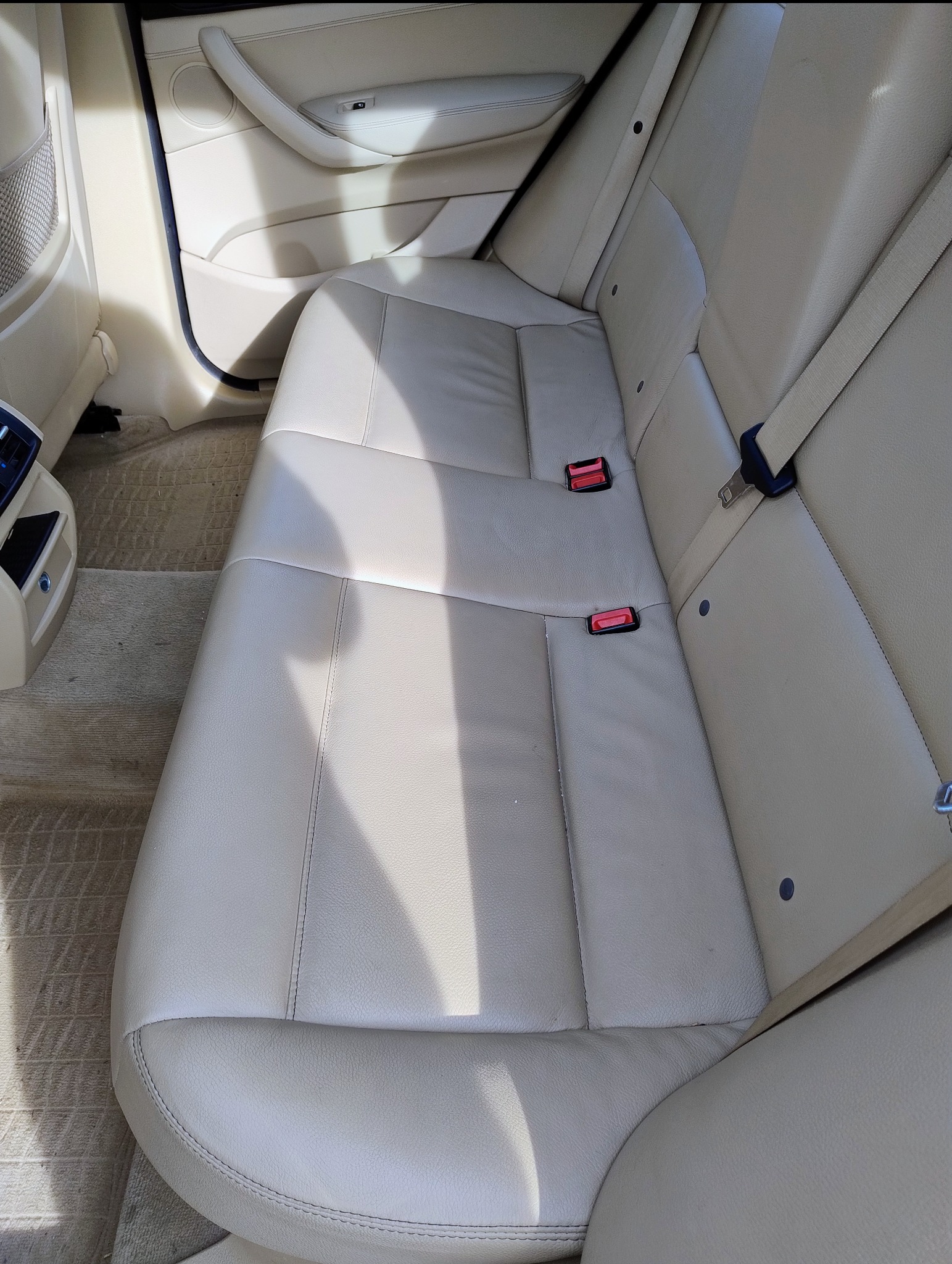 Beżowa tylna kanapa skórzana w samochodzie, widoczne pasy bezpieczeństwa i dywaniki podłogowe.