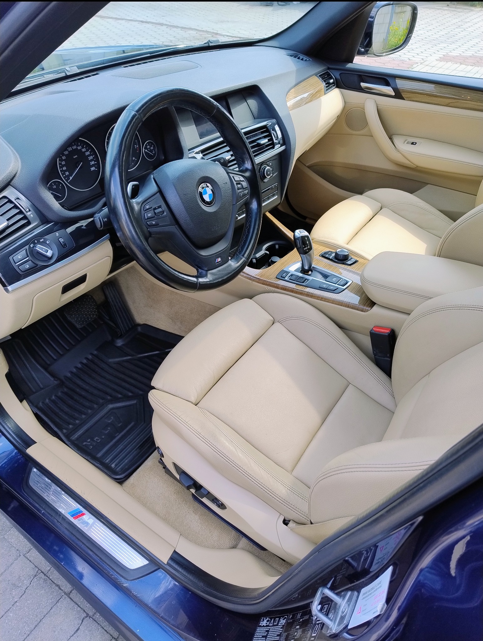 Wnętrze samochodu BMW X3 z jasną skórzaną tapicerką, widoczne czarne gumowe dywaniki z logo 'No. 4' oraz emblemat M Performance na progu.