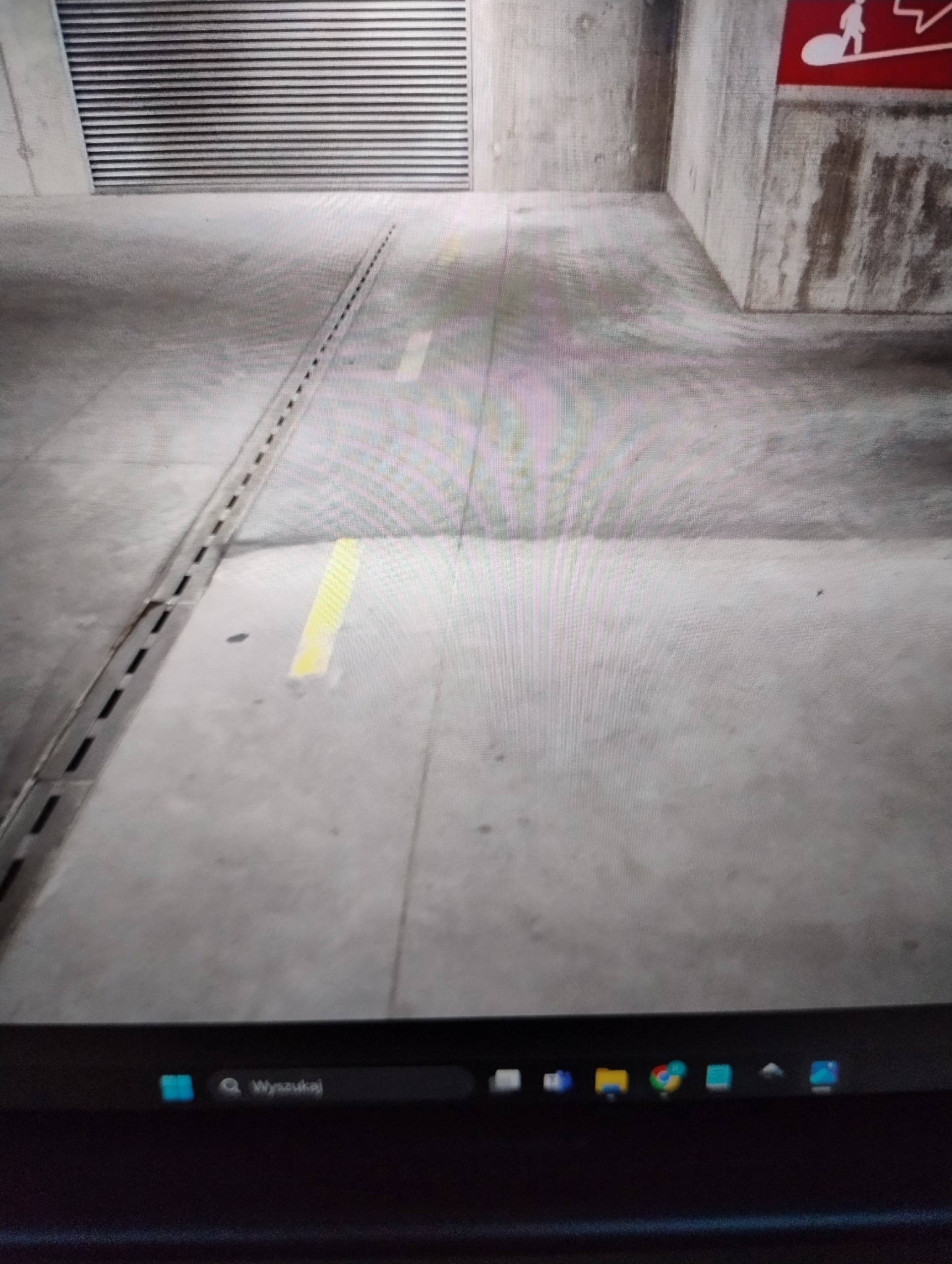 Szare betonowe podłoże w garażu podziemnym z widocznymi liniami i kratką odpływową, fragment ściany z oznaczeniem ewakuacyjnym.