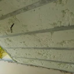 Poddasze z widoczną warstwą izolacji natryskowej między metalowymi profilami, fragment okna z żółtą folią ochronną.