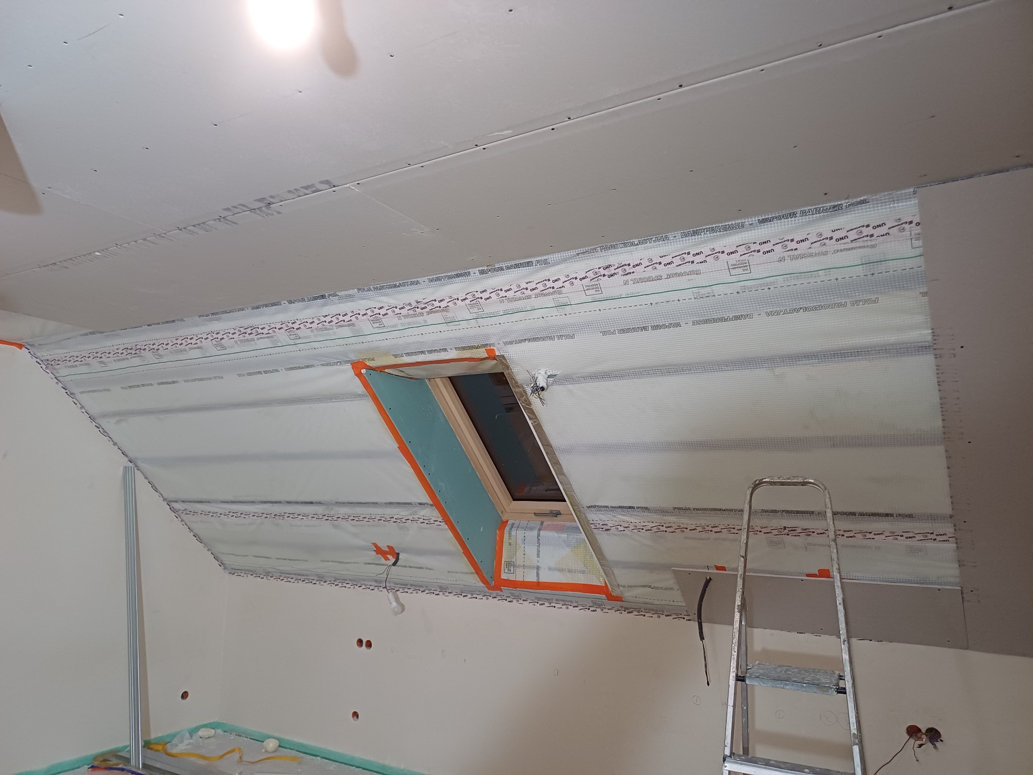 Poddasze w trakcie prac wykończeniowych: widoczna izolacja folią paroizolacyjną, okno dachowe z obróbką i częściowo zamontowane płyty gipsowo-kartonowe na skosach, drabina oparta o ścianę.