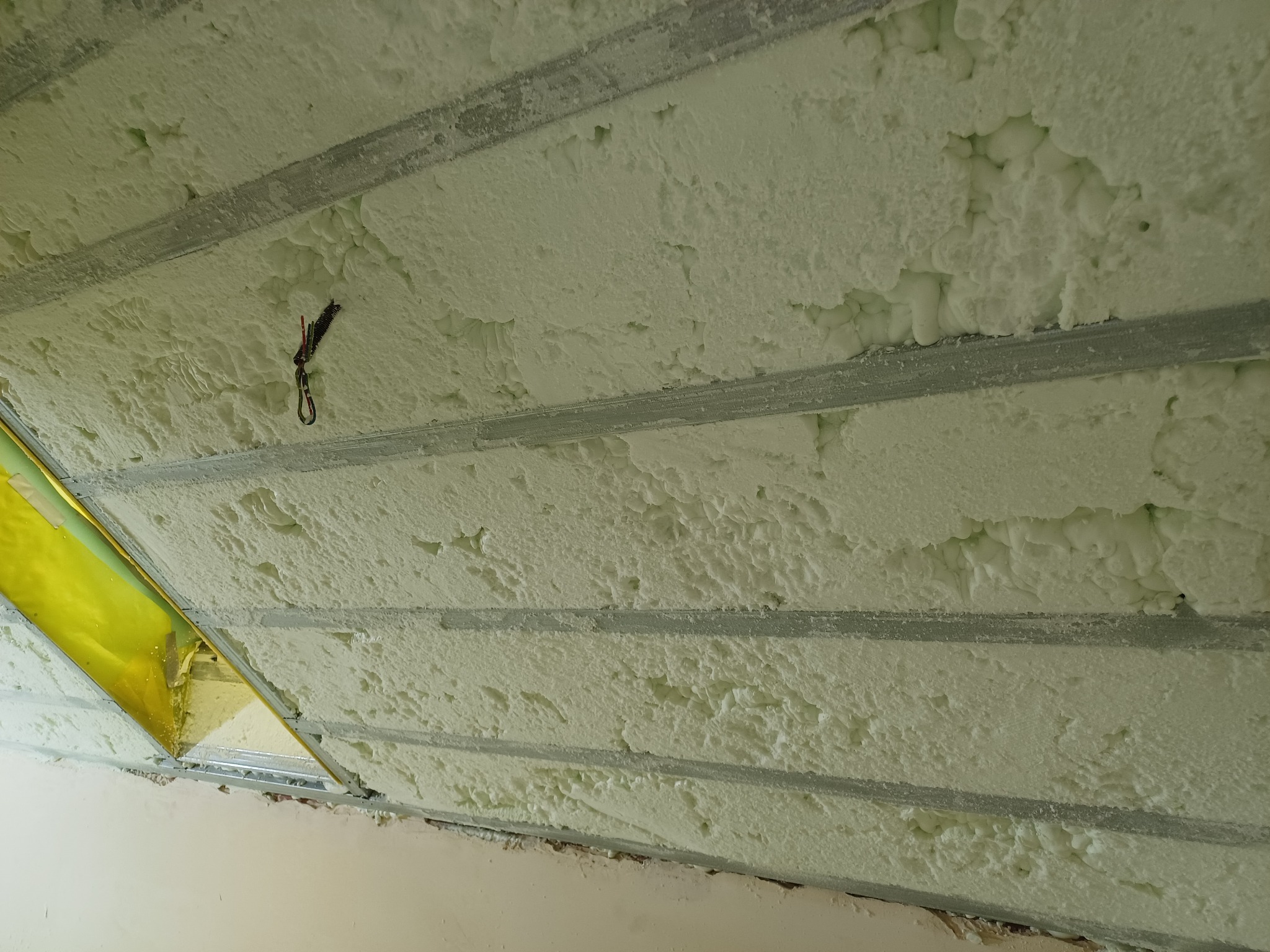 Poddasze z widoczną warstwą izolacji natryskowej między metalowymi profilami, fragment okna z żółtą folią ochronną.