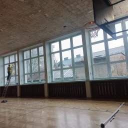 Pro-Dach - Pracownik na drabinie maluje okna w sali gimnastycznej z parkietem i koszem do koszykówki, widoczne stare okna i zniszczony sufit.