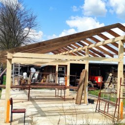 Pro-Dach - Konstrukcja szkieletowa z drewna, prawdopodobnie zadaszenie tarasu lub altany, w trakcie budowy na betonowej wylewce, z widocznym ogrodem i budynkiem w tle.