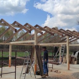 Pro-Dach - Konstrukcja drewniana z elementami dachu w trakcie budowy, widoczni dwaj pracownicy, drabiny i narzędzia, w tle drzewa i błękitne niebo z chmurami.