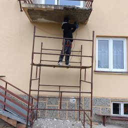 Pro-Dach - Pracownik na rusztowaniu, skuwa uszkodzony betonowy fragment balkonu w budynku o beżowej elewacji, widoczne gruz i odłamki betonu na ziemi.