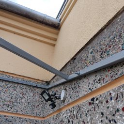 Pro-Dach - Zamontowane oświetlenie zewnętrzne z czujnikiem ruchu na elewacji budynku w Tarnowie z widoczną rynną i konstrukcją wsporczą.