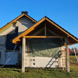 Surowy stan domu w trakcie budowy z widoczną konstrukcją dachu, oknami zabezpieczonymi folią i częściowo otynkowanymi ścianami z betonu komórkowego.