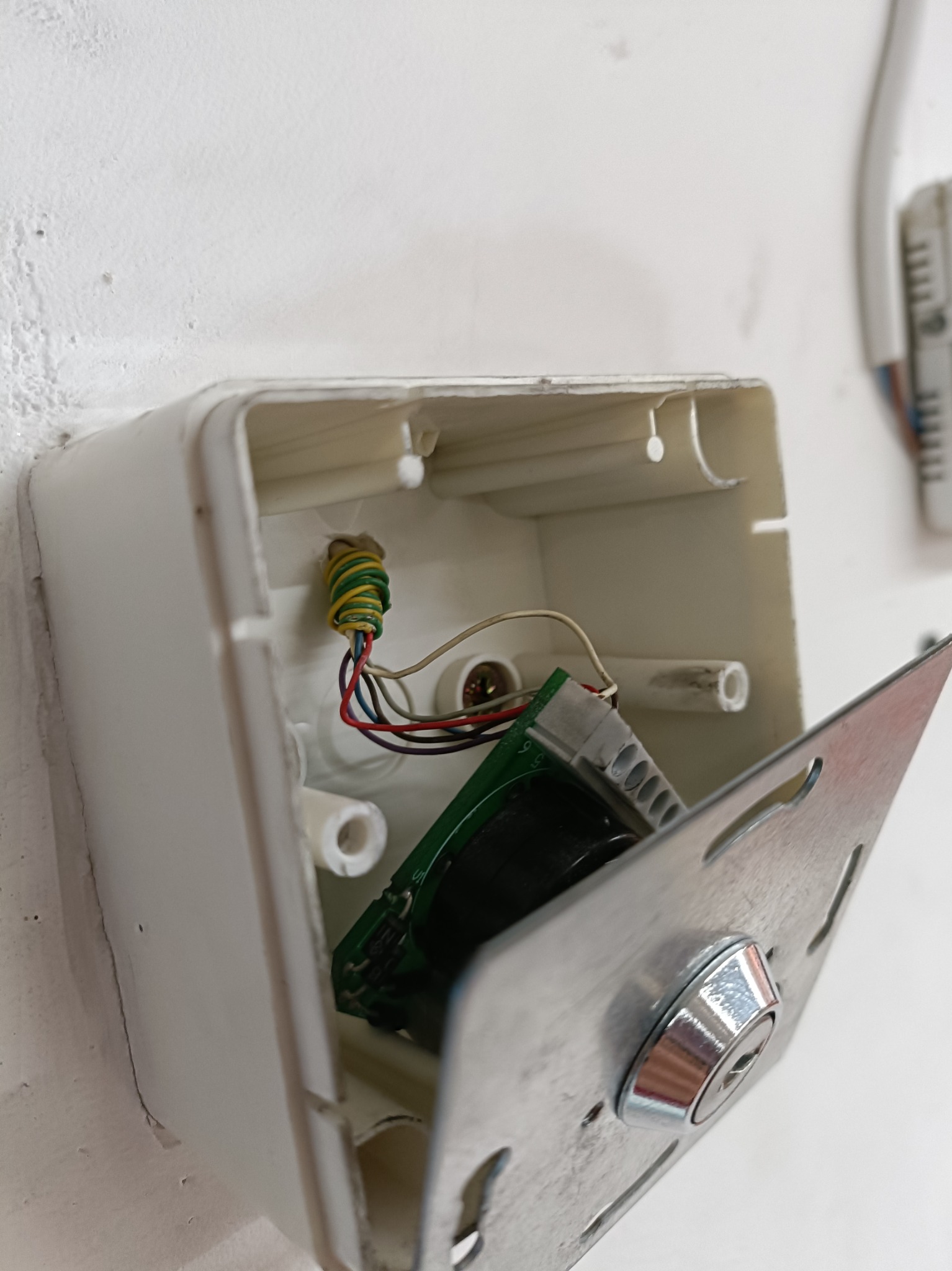 Otwarta biała puszka instalacyjna z widocznymi przewodami zabezpieczonymi zielono-żółtą taśmą, płytką drukowaną i metalową osłoną z zamkiem na białej ścianie.