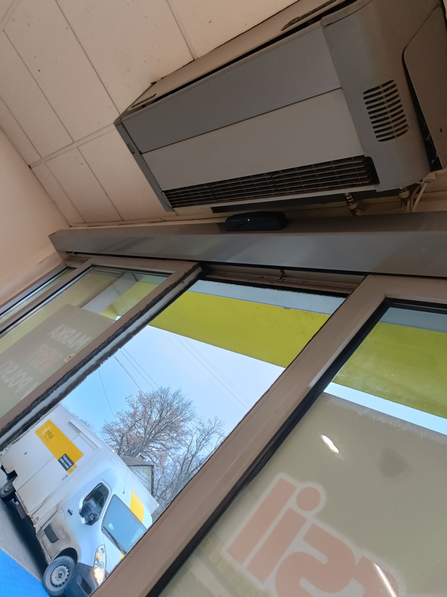 Widok z wnętrza budynku przez okno, na którym widać odbicie białego samochodu dostawczego InPost, na suficie zamontowany grzejnik przemysłowy.