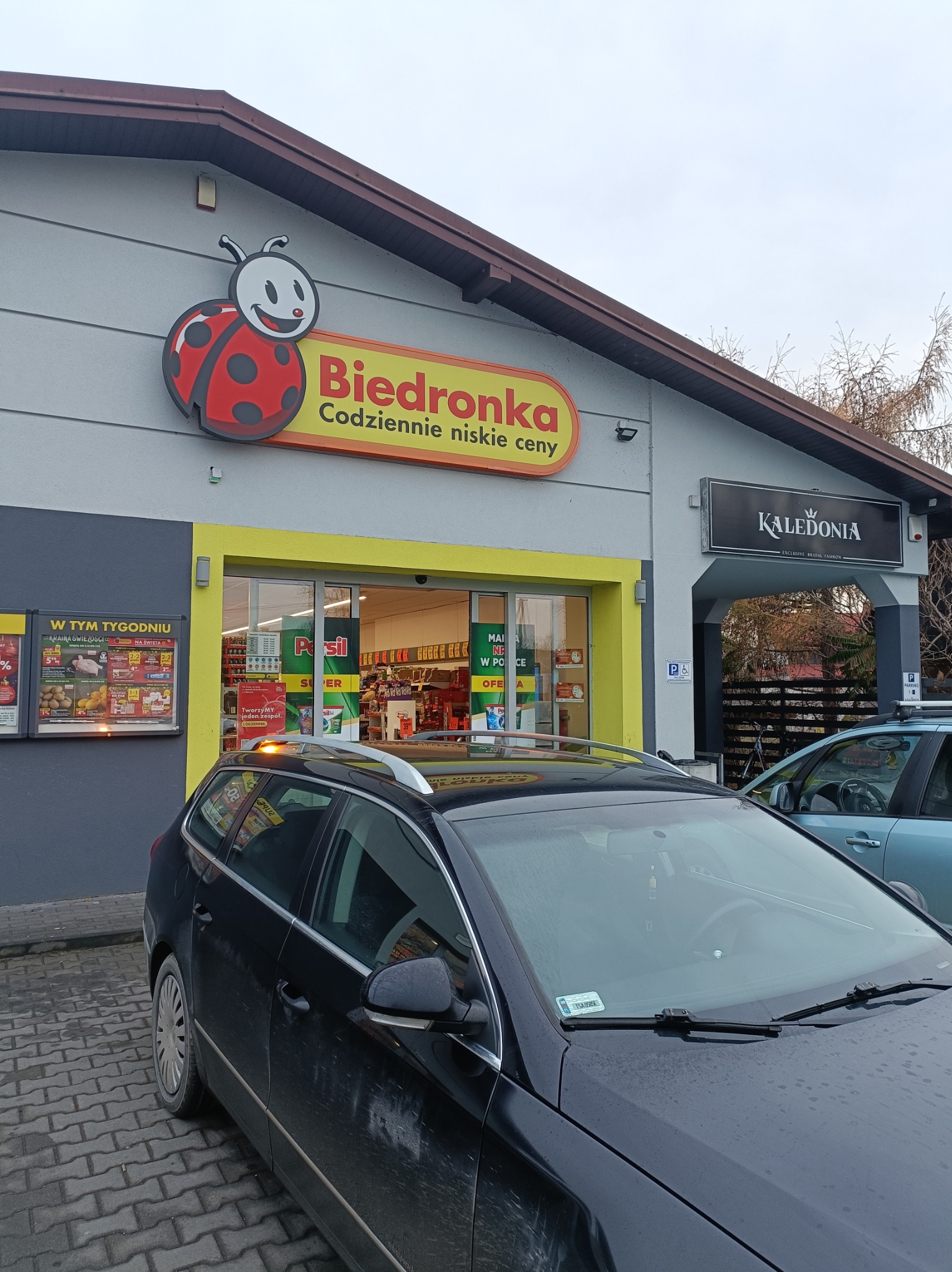 Budynek sklepu Biedronka z charakterystycznym logo biedronki i żółtym obramowaniem wejścia, obok salon sukien ślubnych Kaledonia, zaparkowany czarny samochód na pierwszym planie.