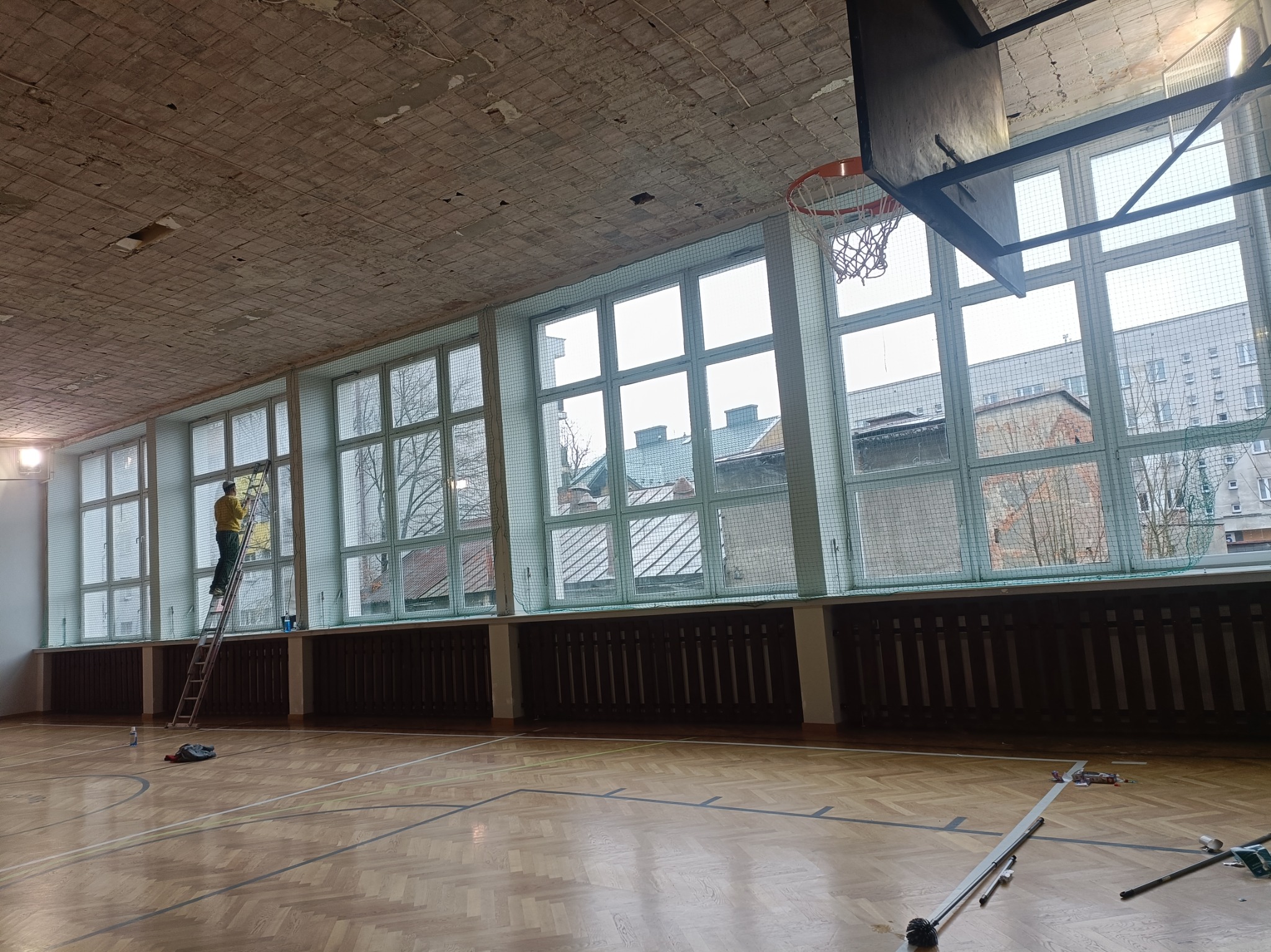 Pracownik na drabinie maluje okna w sali gimnastycznej z parkietem i koszem do koszykówki, widoczne stare okna i zniszczony sufit.