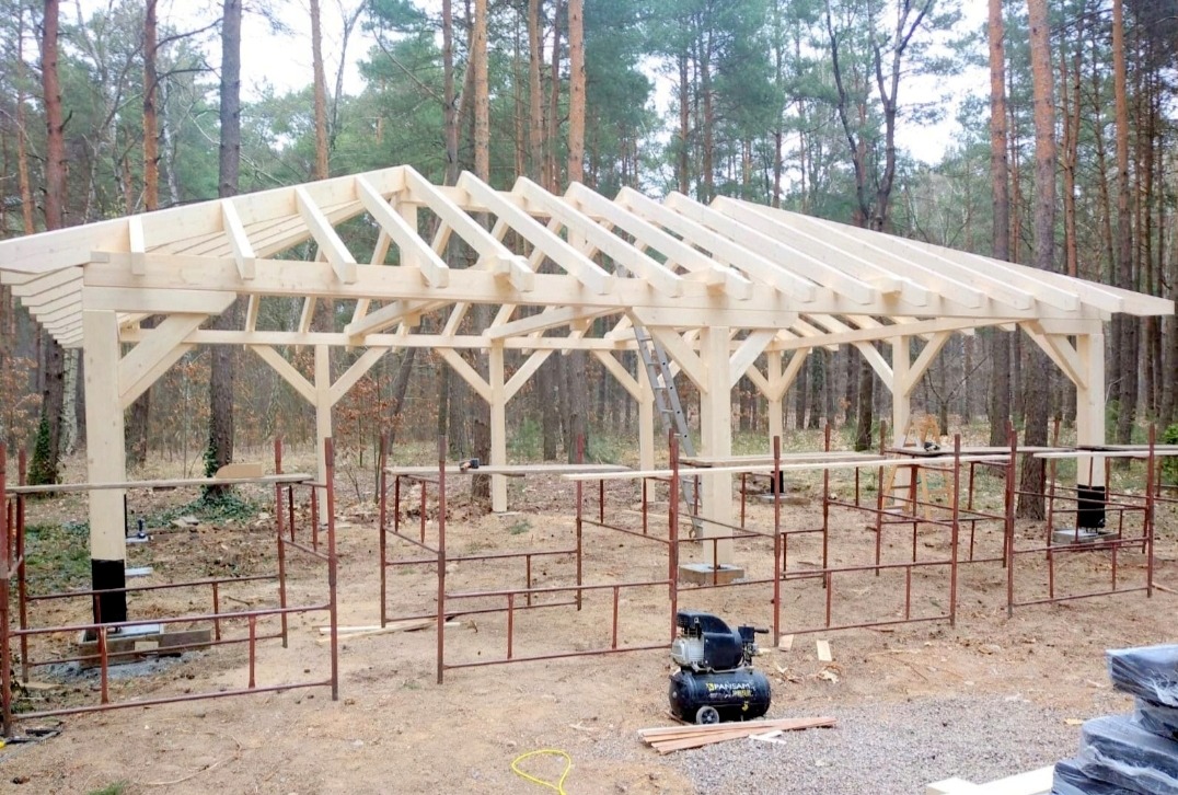 Konstrukcja drewnianej altany w trakcie budowy, widoczne rusztowania i kompresor powietrza, w tle las iglasty.