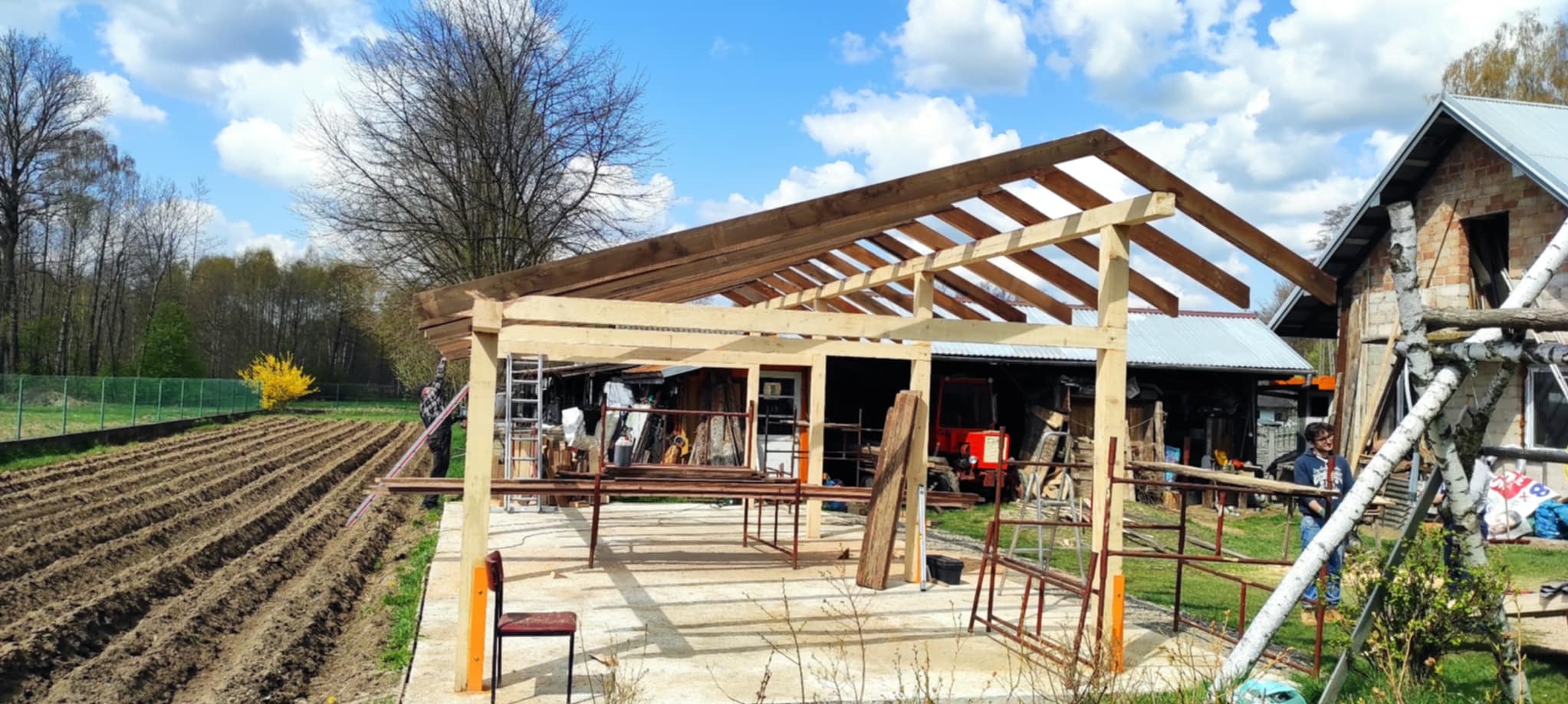 Konstrukcja szkieletowa z drewna, prawdopodobnie zadaszenie tarasu lub altany, w trakcie budowy na betonowej wylewce, z widocznym ogrodem i budynkiem w tle.