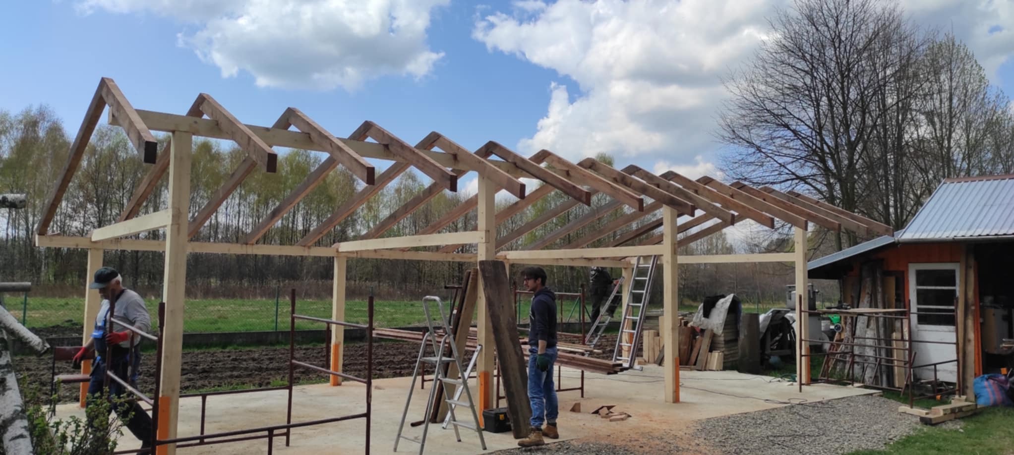 Konstrukcja drewniana z elementami dachu w trakcie budowy, widoczni dwaj pracownicy, drabiny i narzędzia, w tle drzewa i błękitne niebo z chmurami.