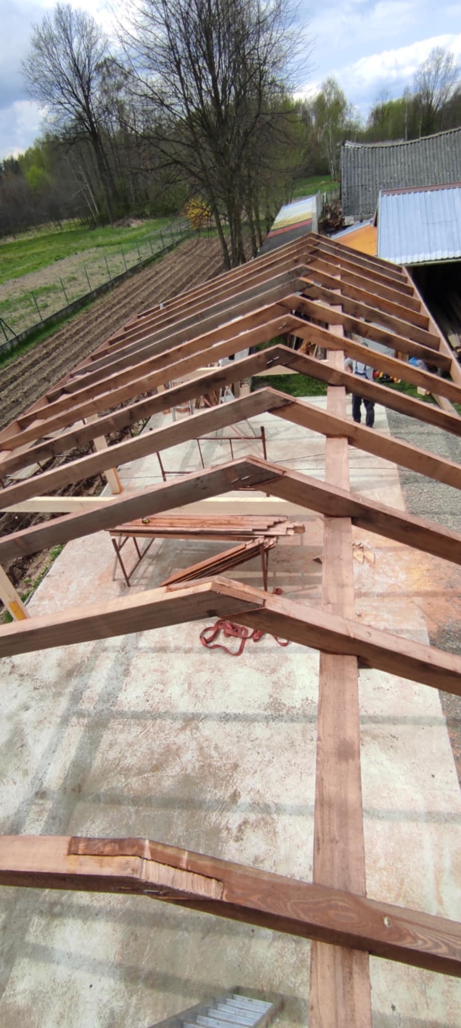 Konstrukcja więźby dachowej z drewnianych belek, widok z góry, w tle pole uprawne i drzewa.