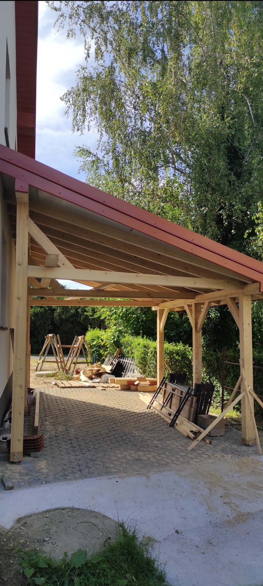Konstrukcja drewnianej wiaty z czerwonym dachem w trakcie budowy, widoczne rusztowania i materiały budowlane na brukowanym podłożu, w tle zieleń drzew.