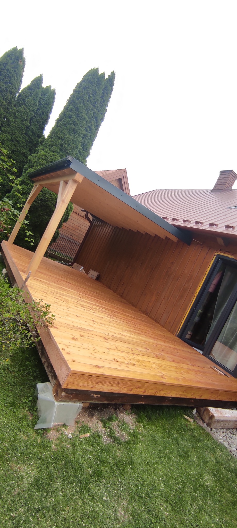 Nowo wybudowany drewniany taras z zadaszeniem przy domu z brązowym dachem, widoczne drzewa iglaste w tle.