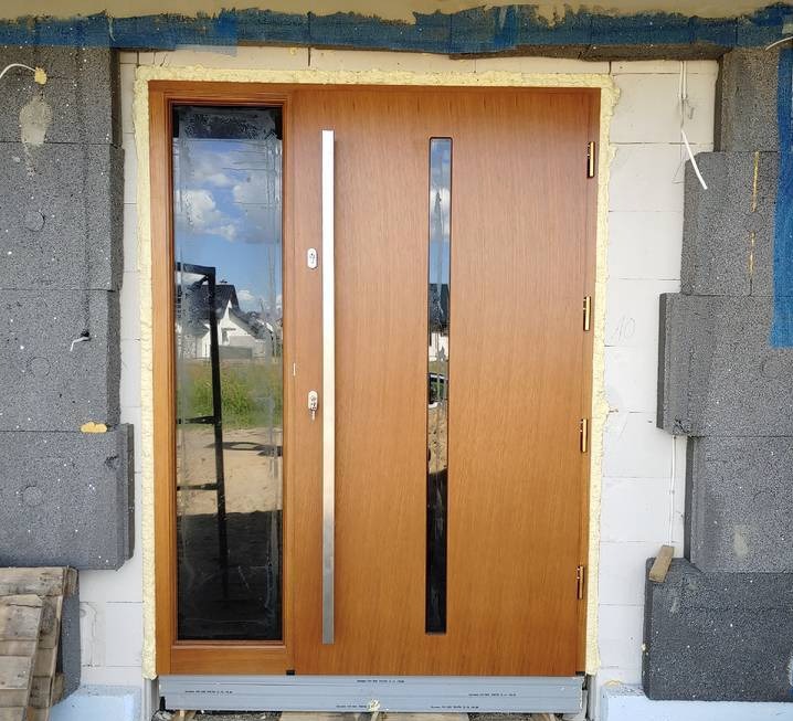 Montaż nowoczesnych drzwi zewnętrznych z długim, pionowym uchwytem i bocznym oknem, widoczne pianowanie uszczelniające ościeżnicę w surowym murze z szarego betonu komórkowego.