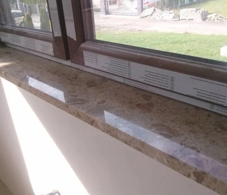 Beżowy marmurowy parapet wewnętrzny z brązową ramą okienną, widok na ogród w tle.