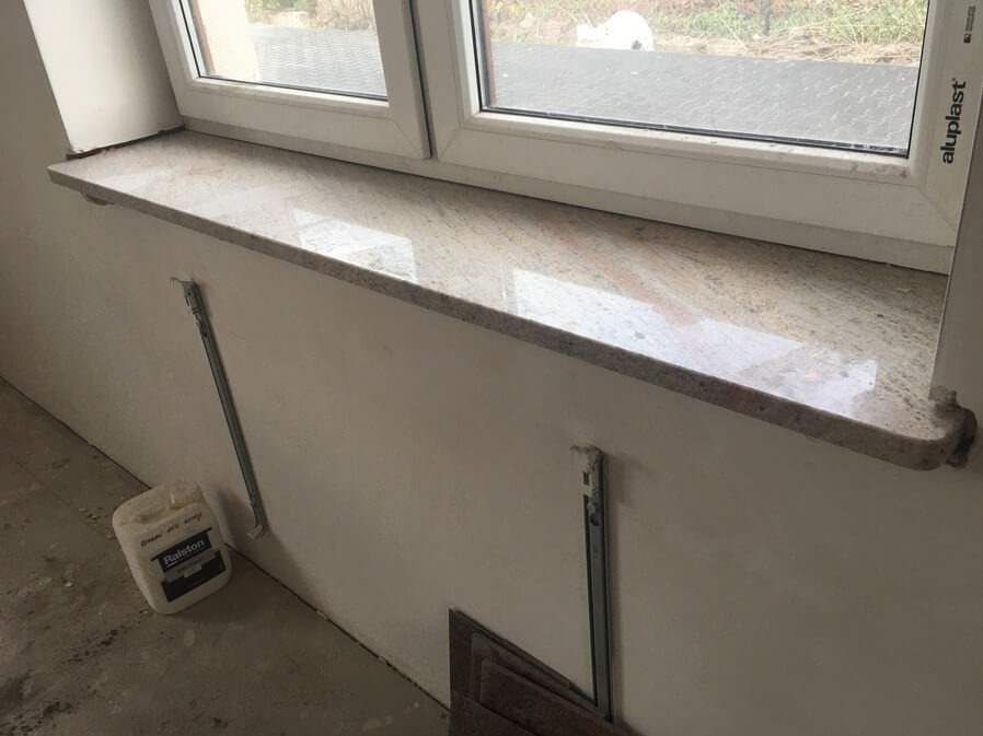 Zamontowany parapet z jasnego, nakrapianego kamienia pod oknem z plastikową ramą, widoczne metalowe wsporniki w ścianie i pojemnik z klejem na podłodze.