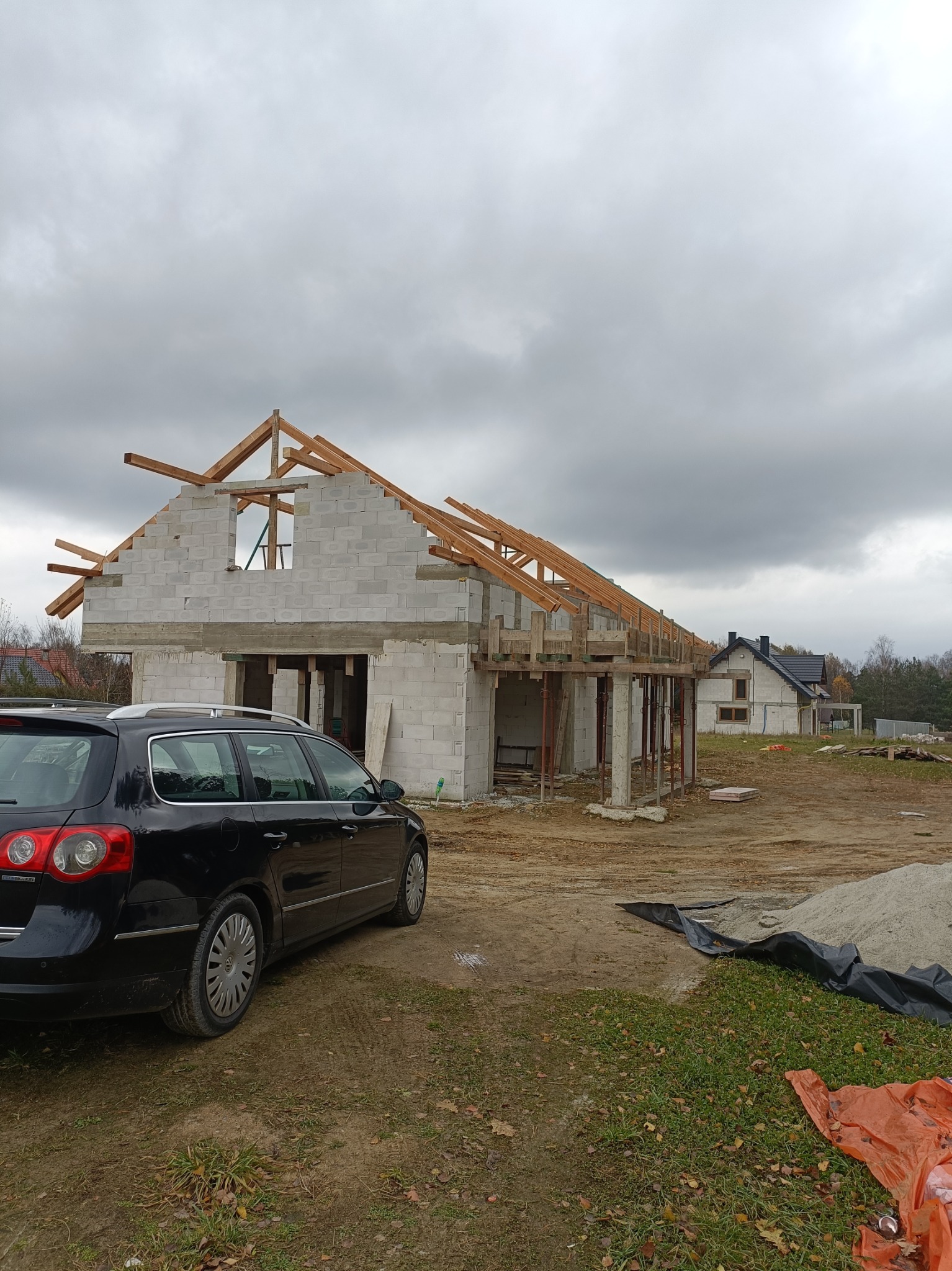 Surowy stan domu w trakcie budowy, widoczne bloczki betonowe, drewniana konstrukcja dachu i szalunki, na pierwszym planie zaparkowany czarny samochód kombi.
