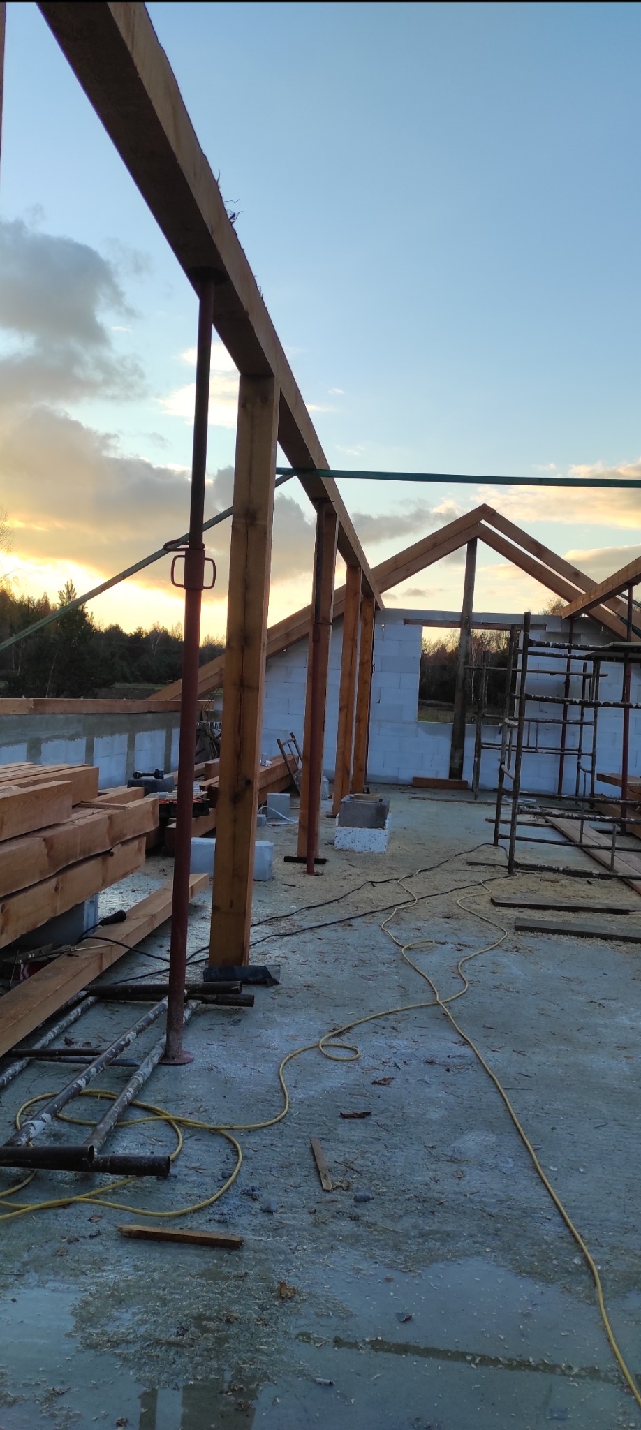 Wznoszenie konstrukcji dachu z drewnianych belek wspartych na stalowych podporach teleskopowych, widoczne bloczki z betonu komórkowego i rusztowanie, tło stanowi zachodzące słońce.