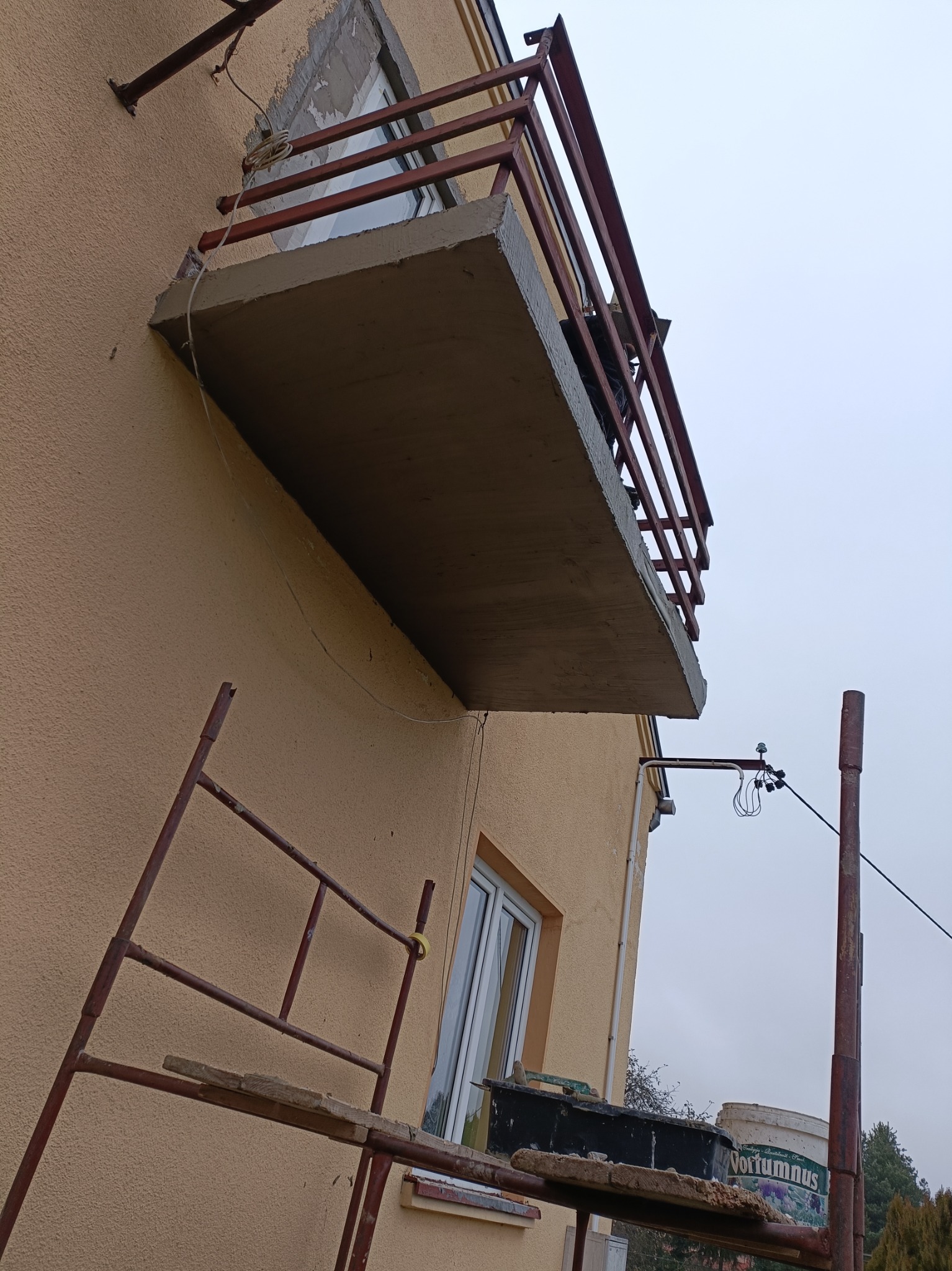 Wzmocniony betonowy balkon z metalową balustradą w trakcie budowy, widoczne rusztowanie i ślady prac murarskich przy oknie, elewacja w kolorze beżowym.