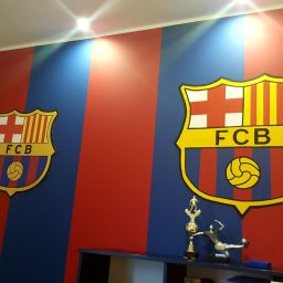 Jakub Kozerski - Pomieszczenie z pionowymi pasami w kolorach czerwonym i niebieskim, ozdobione dwoma herbami FC Barcelona, podświetlone punktowym oświetleniem sufitowym, z widoczną półką i trofeum sportowym.