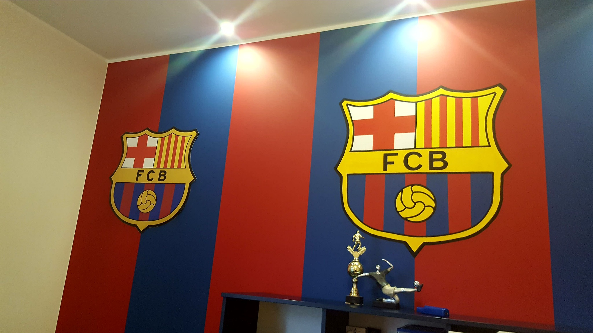 Pomieszczenie z pionowymi pasami w kolorach czerwonym i niebieskim, ozdobione dwoma herbami FC Barcelona, podświetlone punktowym oświetleniem sufitowym, z widoczną półką i trofeum sportowym.