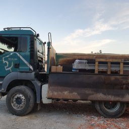 MIDOR-TRANS ADAM MIDOR - Zielony samochód ciężarowy MAN TGS 26.400 z ładunkiem zabezpieczonym folią i drewnianymi belkami, stojący na żwirowym podłożu w słoneczny dzień.