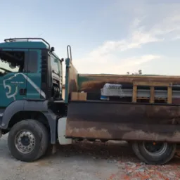 Zielony samochód ciężarowy MAN TGS 26.400 z ładunkiem zabezpieczonym folią i drewnianymi belkami, stojący na żwirowym podłożu w słoneczny dzień.