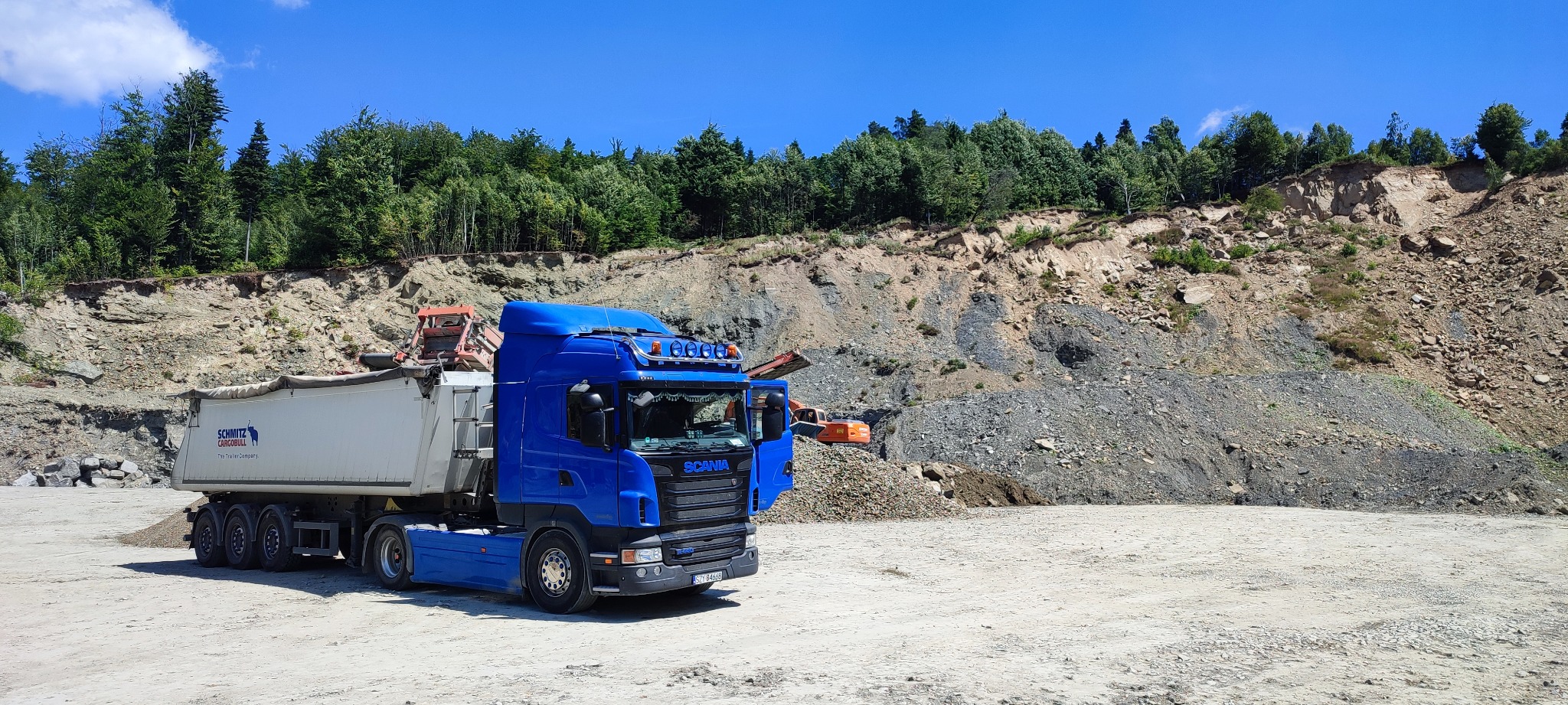 Niebieska ciężarówka Scania z naczepą Schmitz Cargobull na tle kamieniołomu z drzewami i koparką w tle.