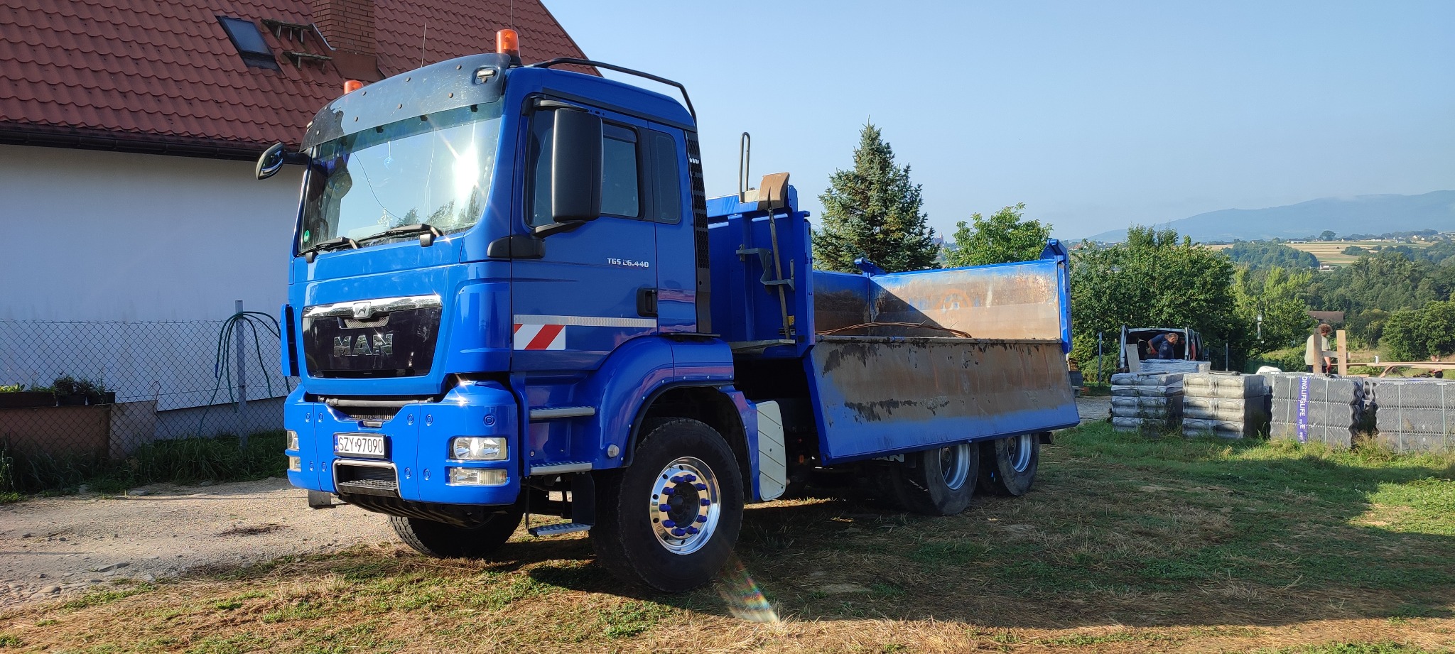 Niebieska ciężarówka MAN TGS 26.440 z wywrotką, zaparkowana na trawie, w tle dom z czerwoną dachówką, drzewa i pagórkowaty krajobraz. W tle widoczne materiały budowlane i osoby pracujące.
