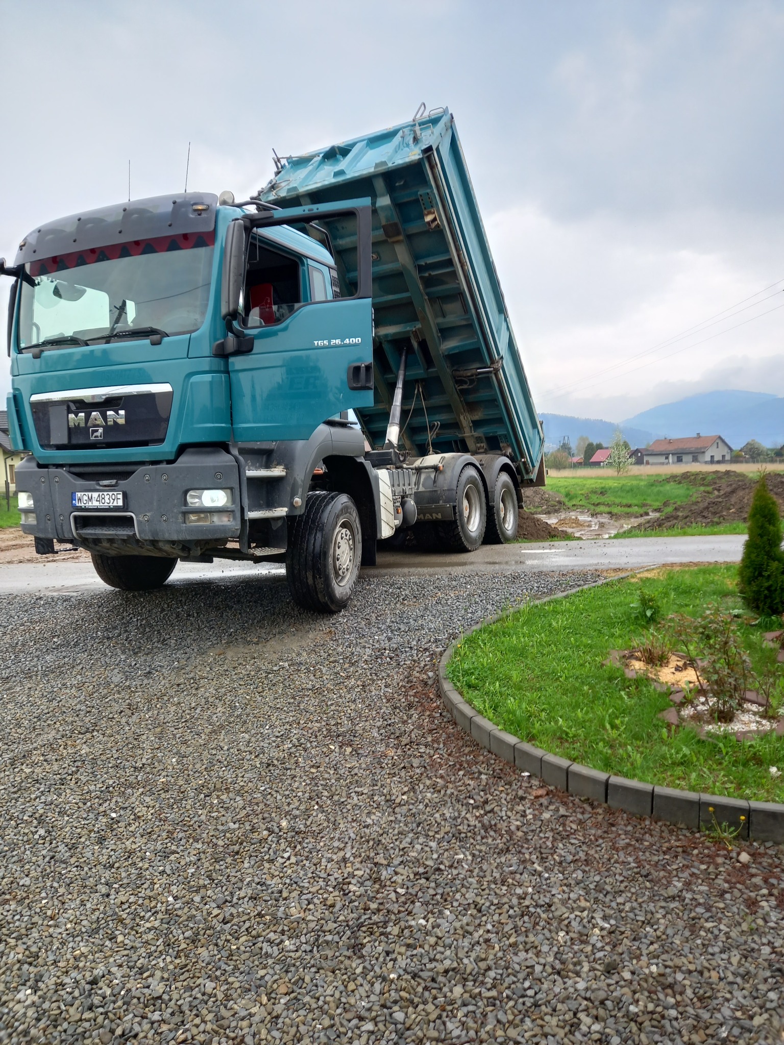 Ciężarówka MAN z podniesioną skrzynią ładunkową wysypująca materiał na żwirową drogę, w tle widoczny krajobraz wiejski z domami i wzgórzami.