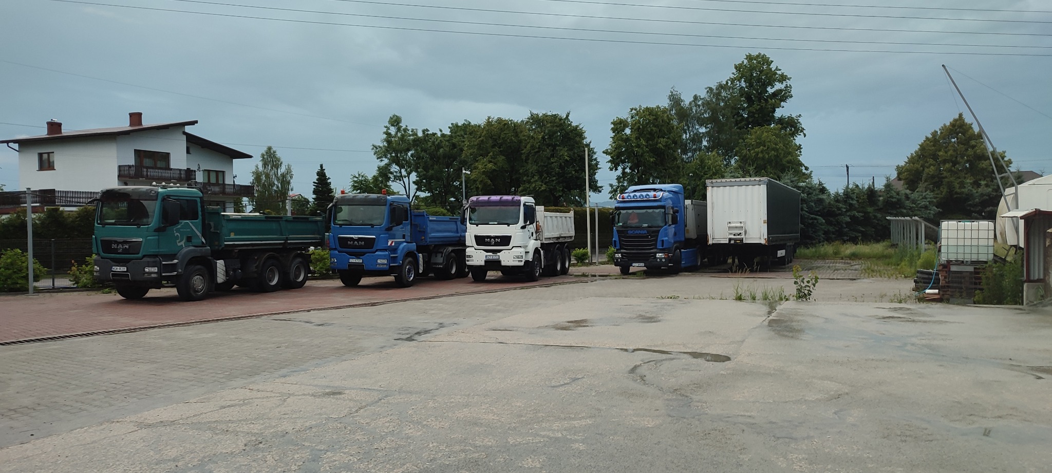 Rząd ciężarówek MAN i Scania, w tym wywrotki, zaparkowanych na betonowym placu, z budynkiem mieszkalnym i drzewami w tle, pochmurne niebo.