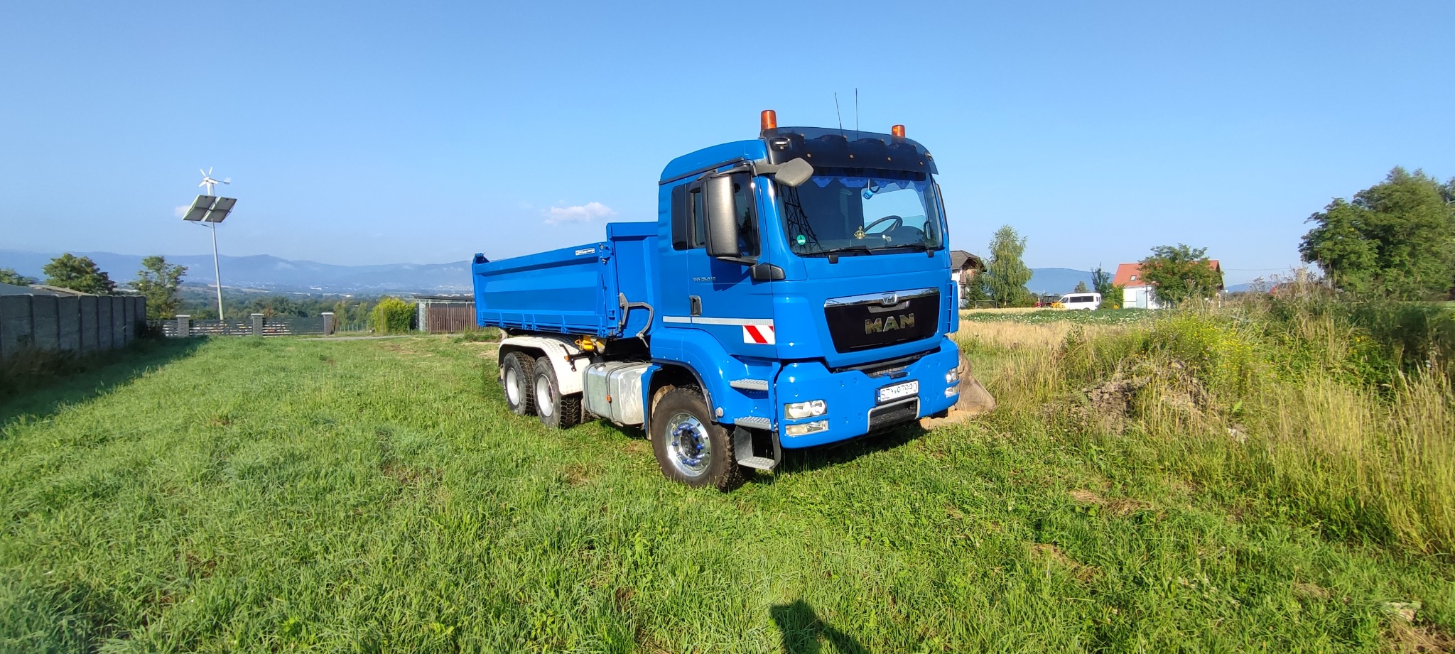 Niebieska wywrotka MAN na zielonym trawiastym terenie, w tle widoczny krajobraz z domami i wzgórzami pod błękitnym niebem.
