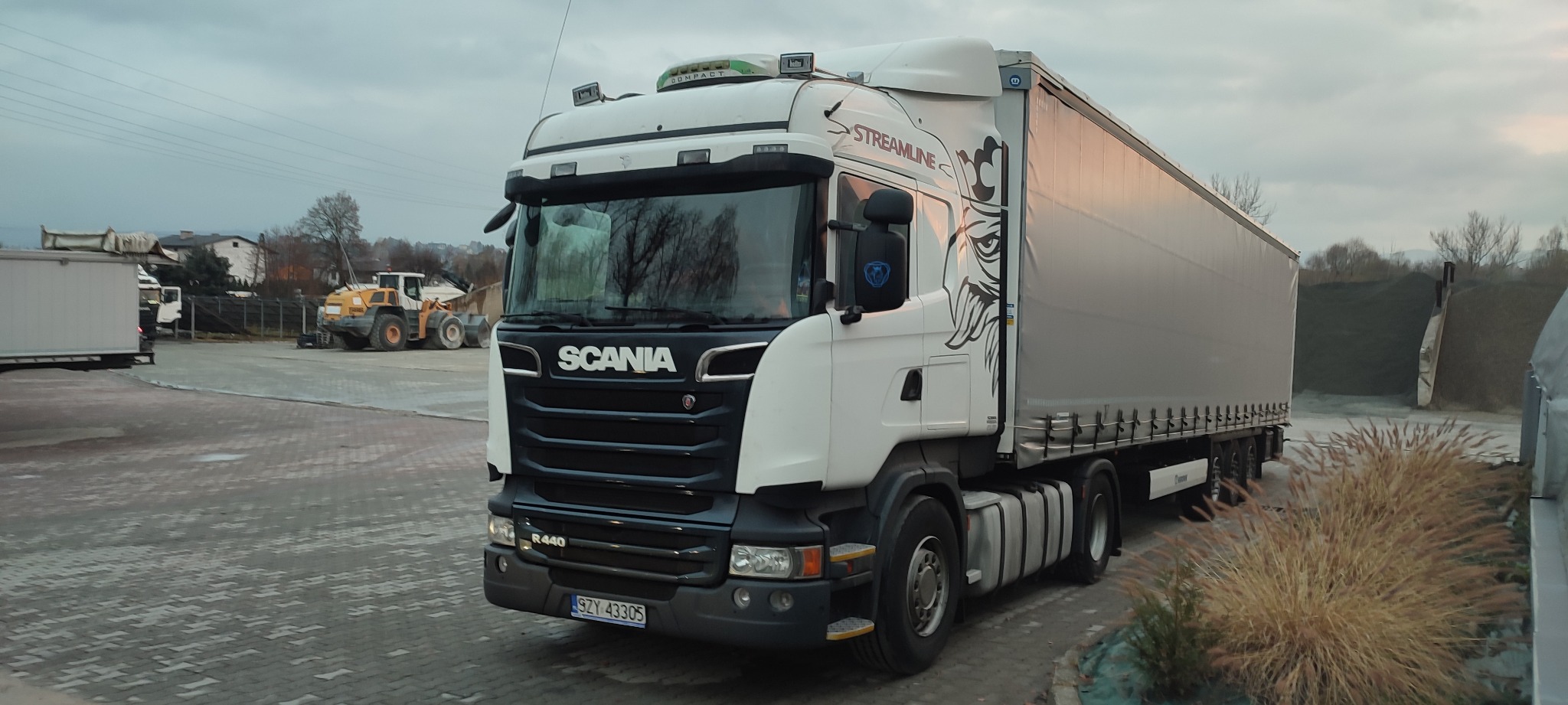 Biała ciężarówka Scania Streamline R440 z naczepą na placu, w tle widoczna ładowarka kołowa oraz hałda ziemi.