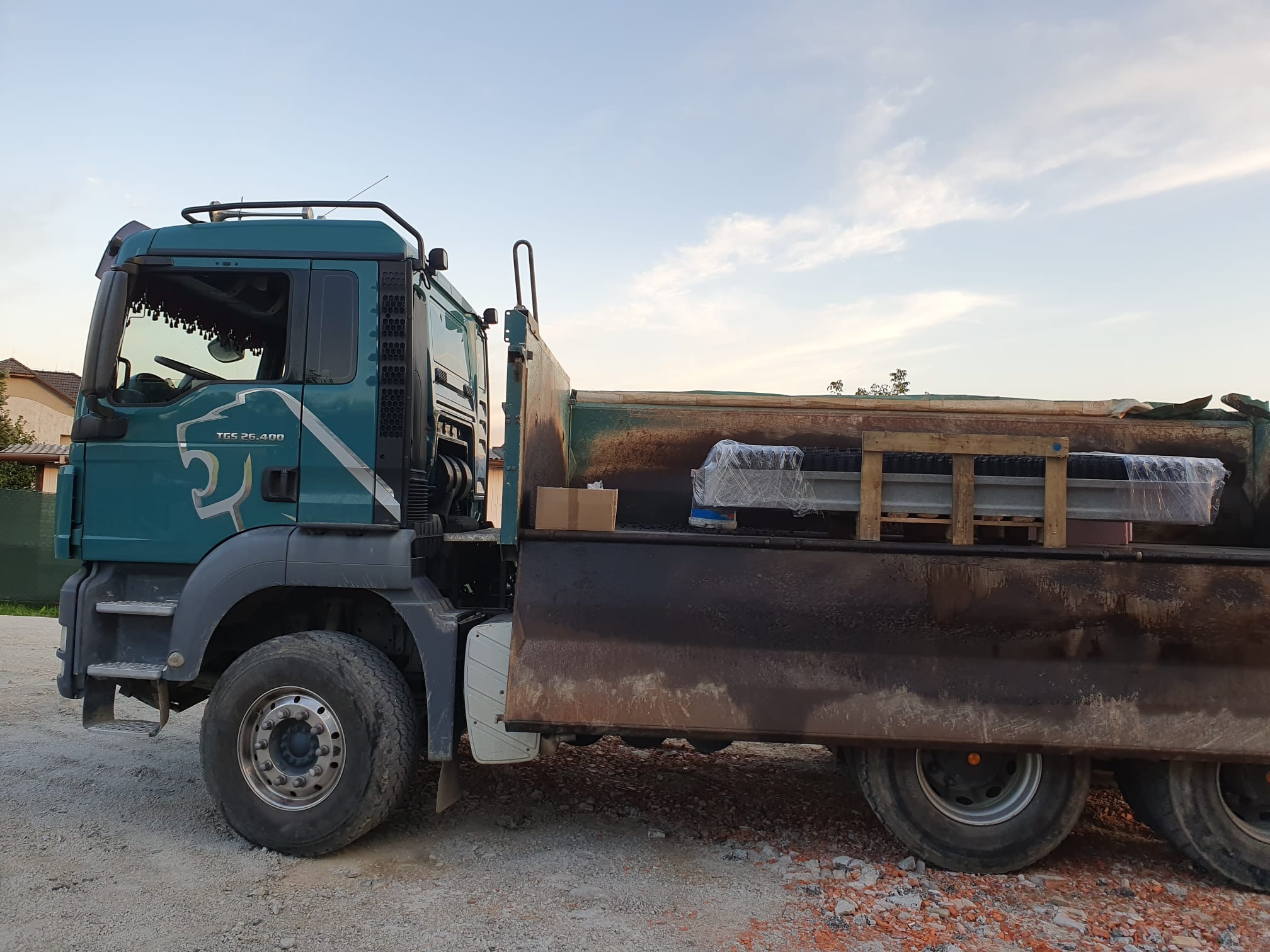 Zielony samochód ciężarowy MAN TGS 26.400 z ładunkiem zabezpieczonym folią i drewnianymi belkami, stojący na żwirowym podłożu w słoneczny dzień.