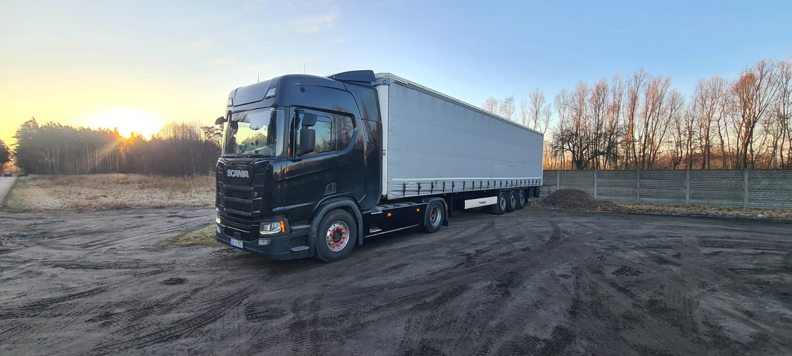 Czarny ciągnik siodłowy Scania z naczepą na błotnistym placu, widoczne ślady opon, w tle betonowy płot i drzewa oświetlone zachodzącym słońcem.