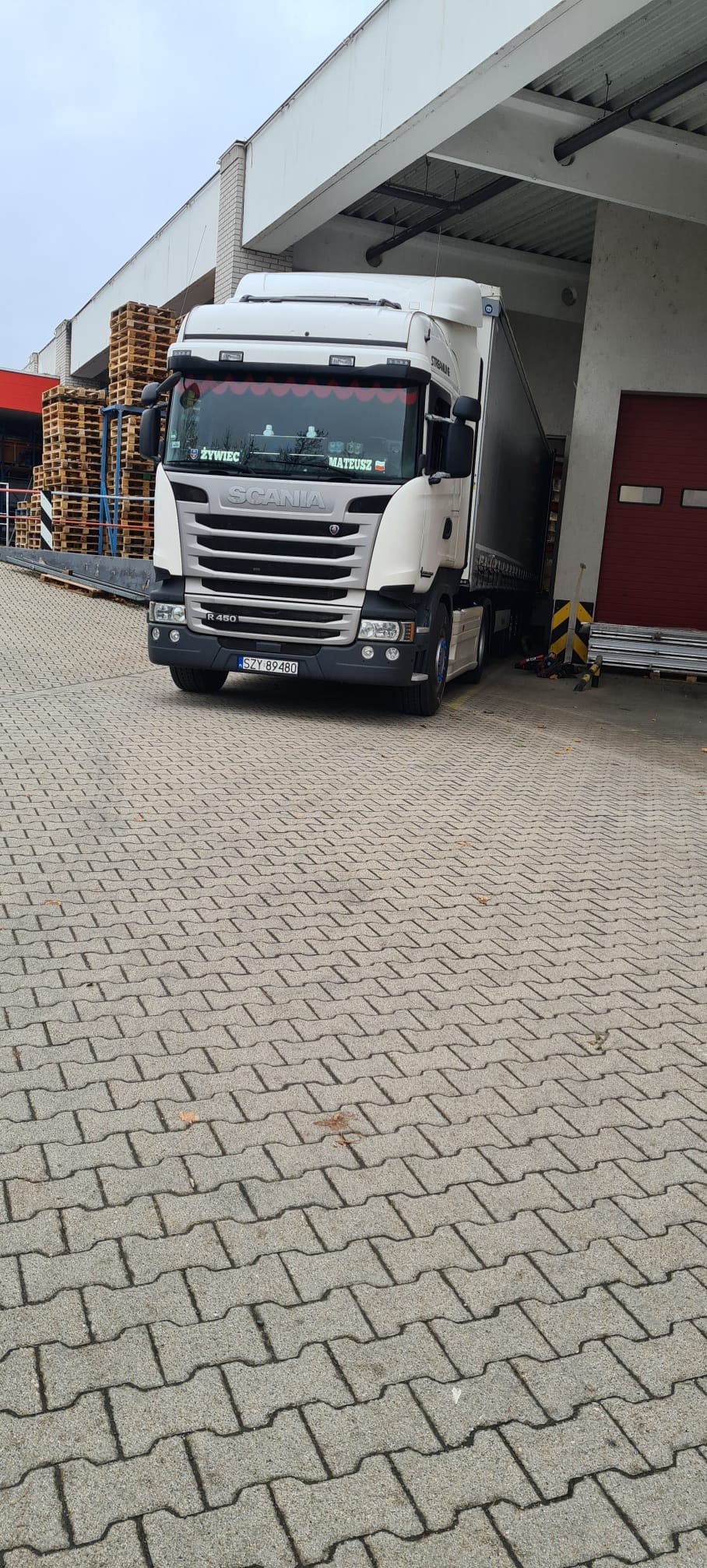 Ciężarówka Scania R450 z naczepą zaparkowana na kostce brukowej w Zarzeczu, przed halą magazynową z rampą załadunkową i stertą palet w tle.