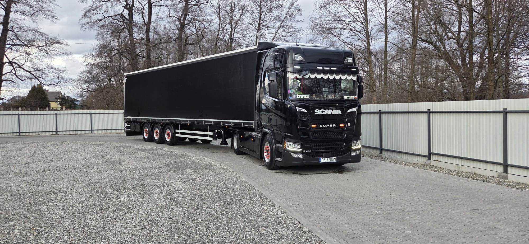 Czarna ciężarówka Scania R450 z naczepą na placu, widok z boku. Czerwone akcenty na kołach. W tle drzewa i ogrodzenie z blachy trapezowej.